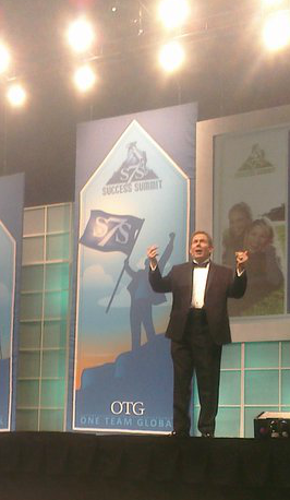 Terry L. Fossum on a large branded stage with banners behind him and stage lights overhead